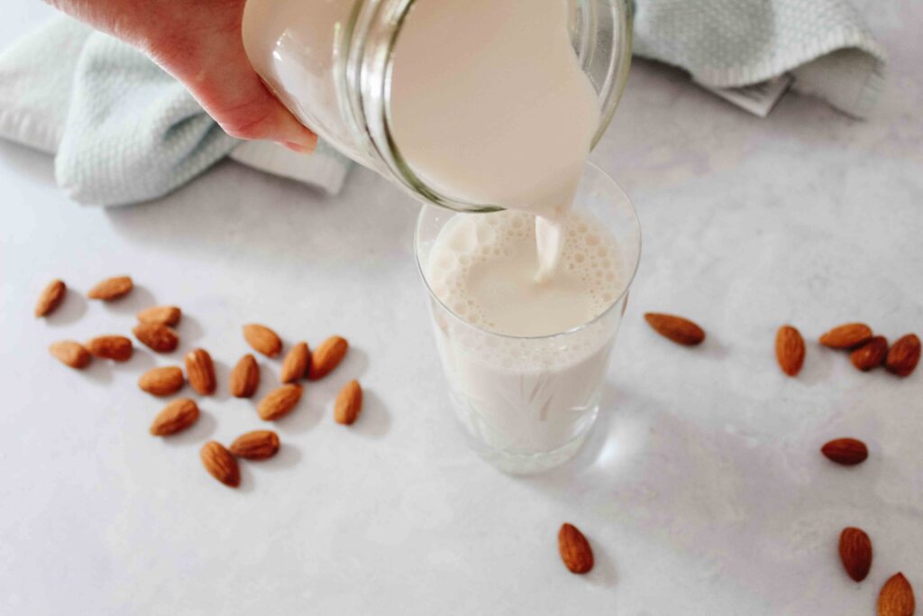 almond milk being poured from mason jar into crystal glass over counter with almonds scattered