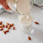 almond milk being poured from mason jar into crystal glass over counter with almonds scattered