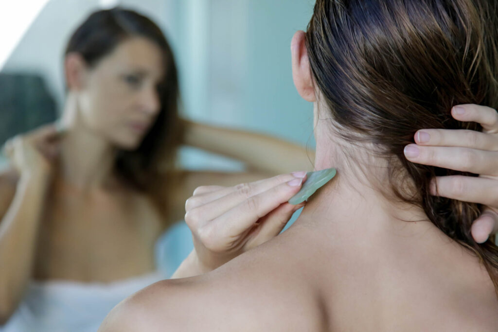 Woman doing Gua Sha facial massage on neck while looking in mirror