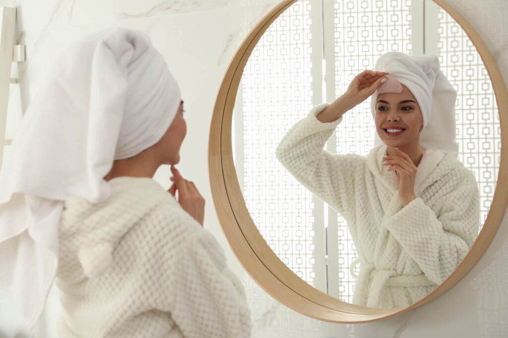 woman doing a facial gua sha massage wearing white robe and towel in hair while looking in mirror