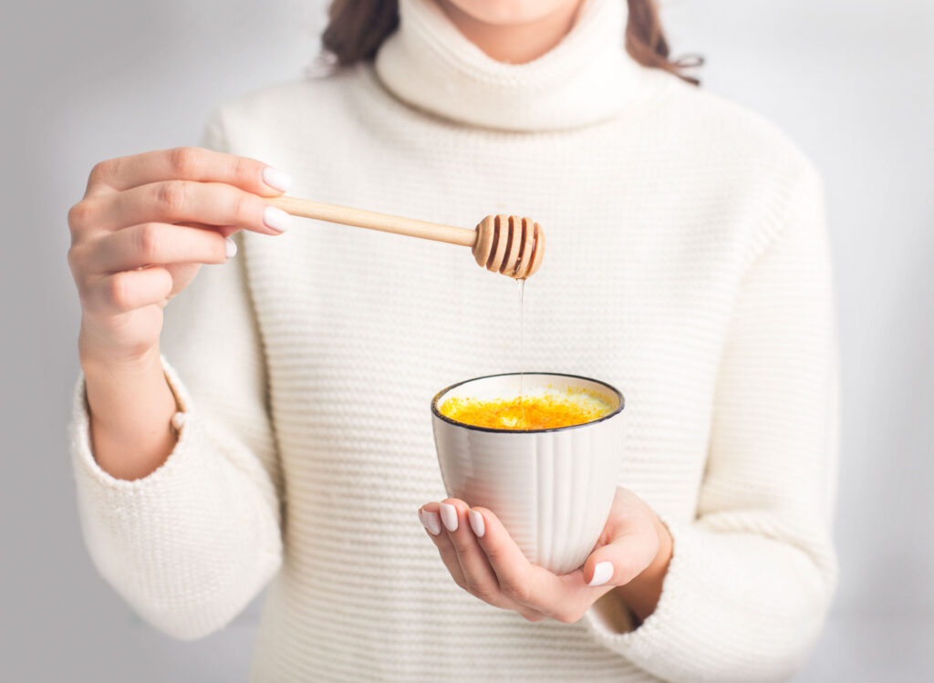 Woman in white sweater holding a cup of turmeric golden milk and holding a honey scoop over cup