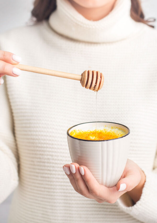 Woman in white sweater holding a cup of turmeric golden milk and holding a honey scoop over cup