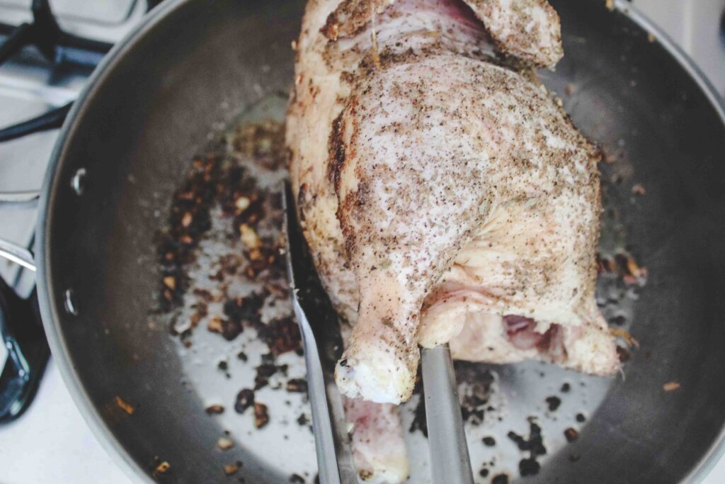 tongs holding whole chicken covered in seasoning being fried in a stainless steel pan