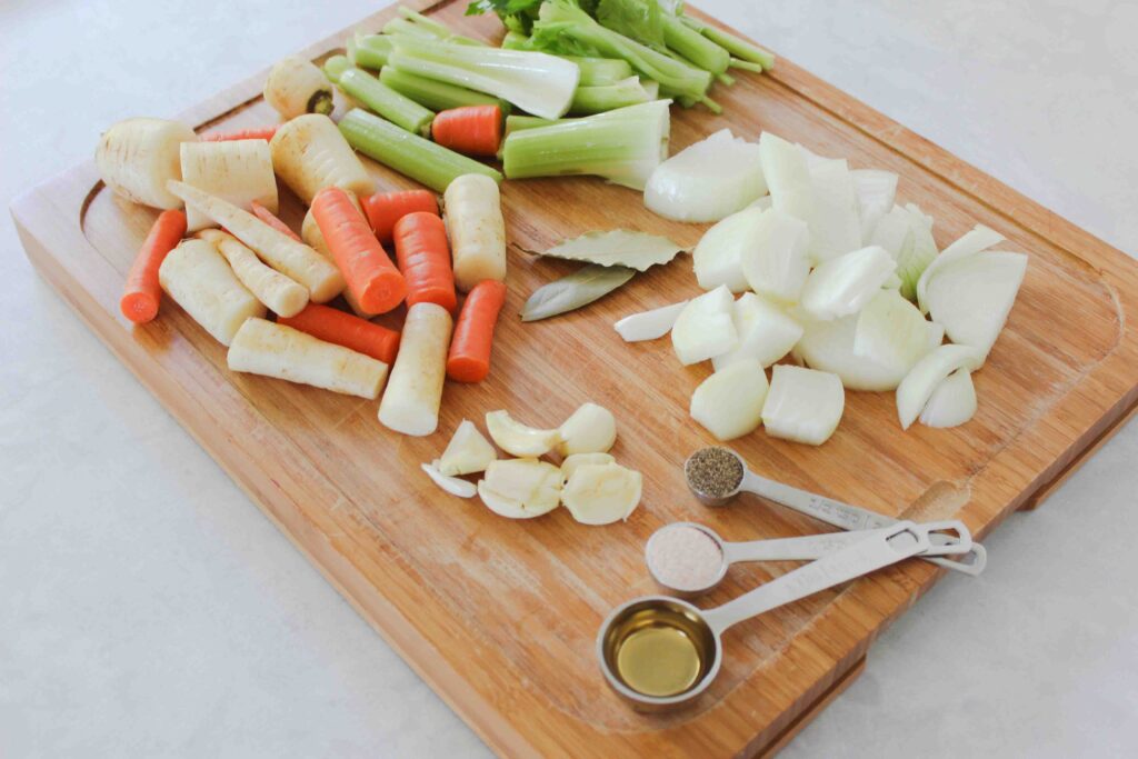 wooden cutting board with chopped celery, carrots and onions, smashed garlic cloves and two bay leaves with tablespoons and teaspoons of seasonings on white counter