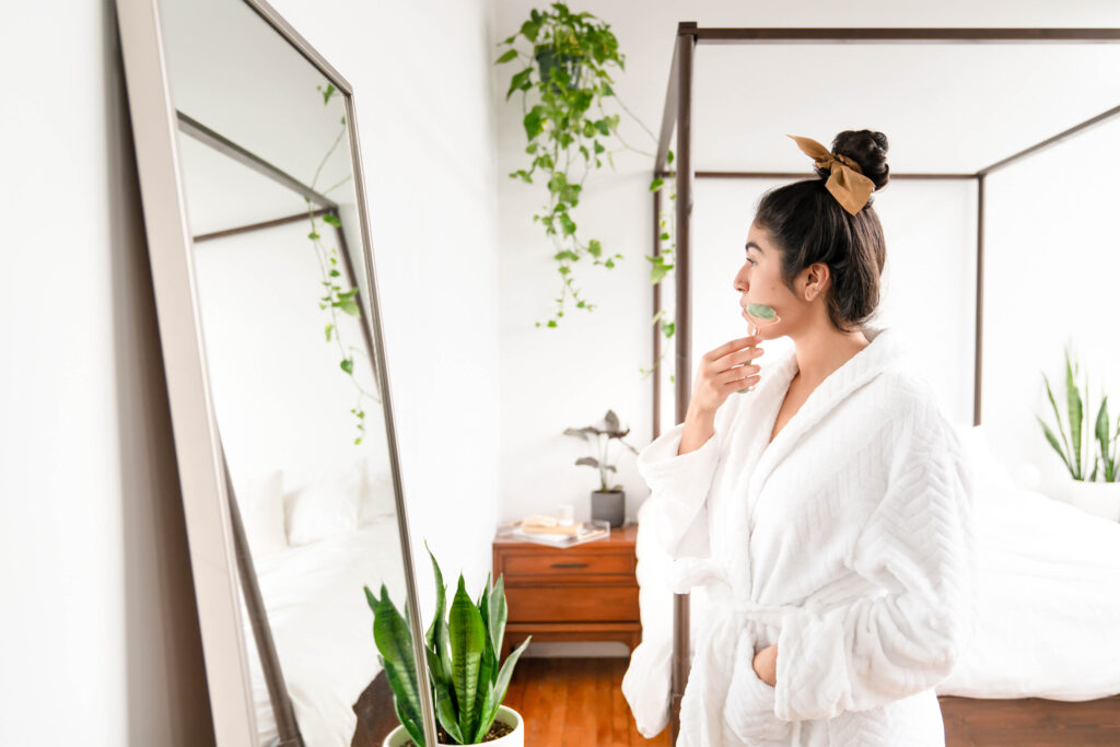 Woman in white robe looking in mirror using jade roller