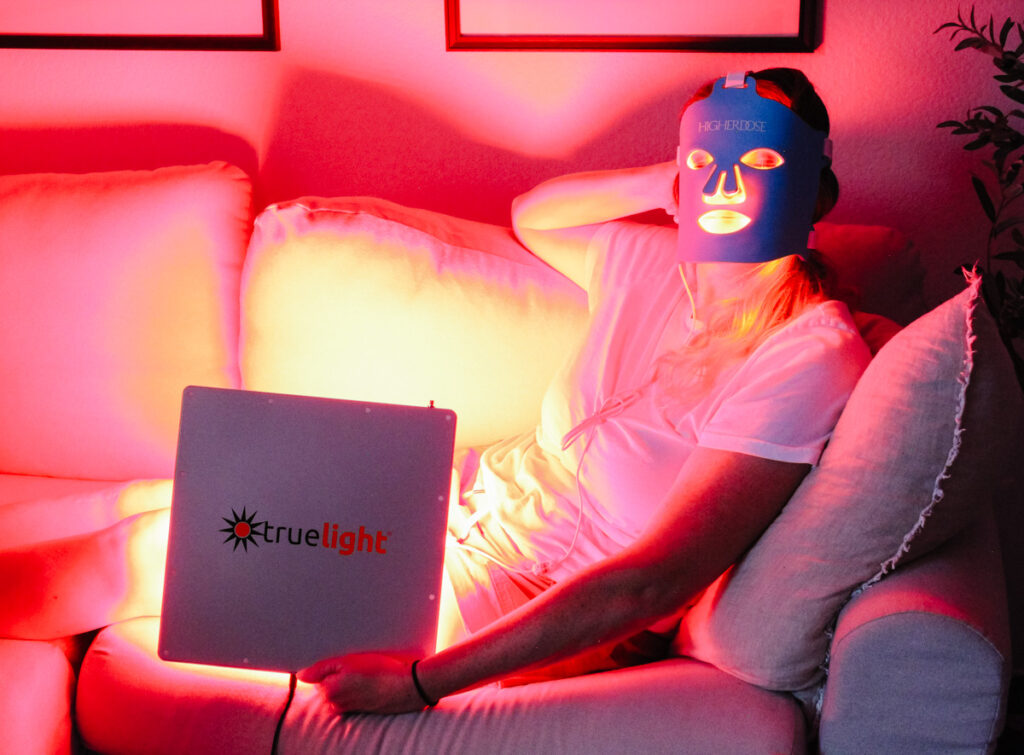 woman on couch holding a TrueLight Energy Square and wearing a Higher Dose Red Light face mask device