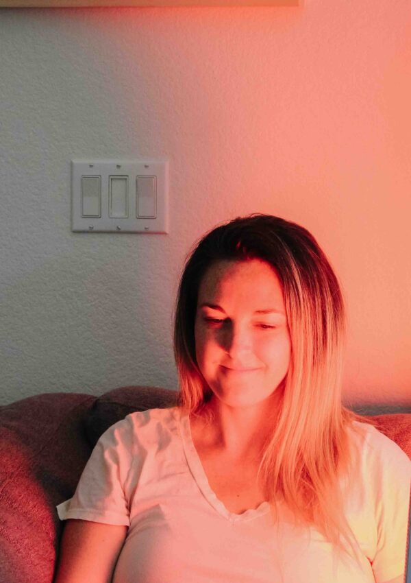 Woman holding TrueLight red light therapy device pointed to her face while sitting on a gray couch