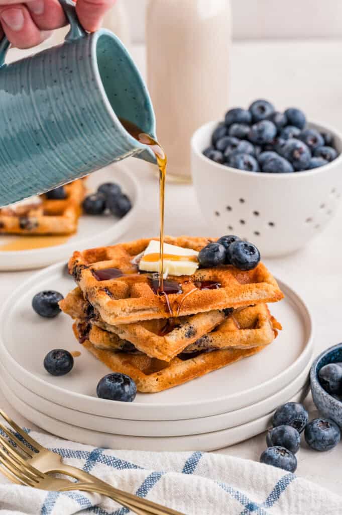 Gluten Free Blueberry Waffles on a plate with butter on top and blueberries on the plate with a hand pouring a cup of maple syrup on the waffles.