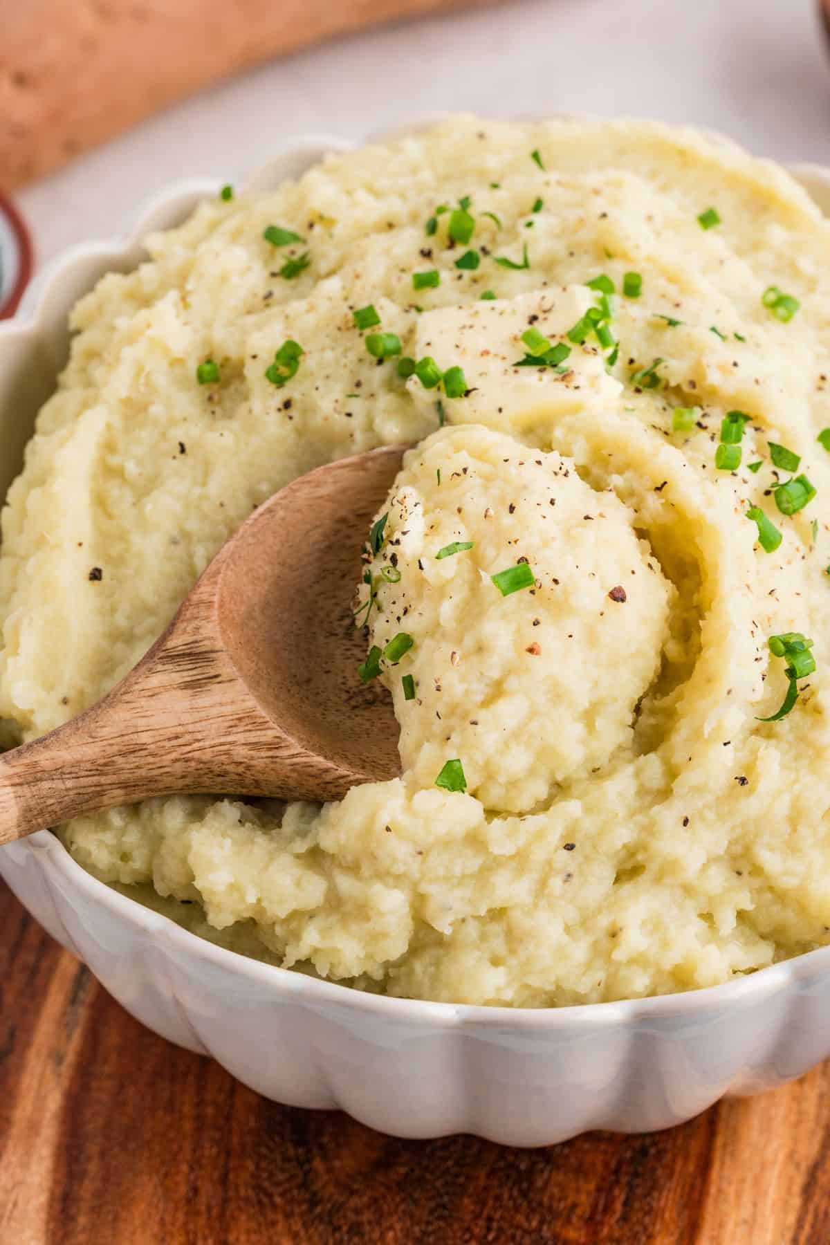 Mashed white sweet potatoes being scooped out of serving dish with a wooden spoon.