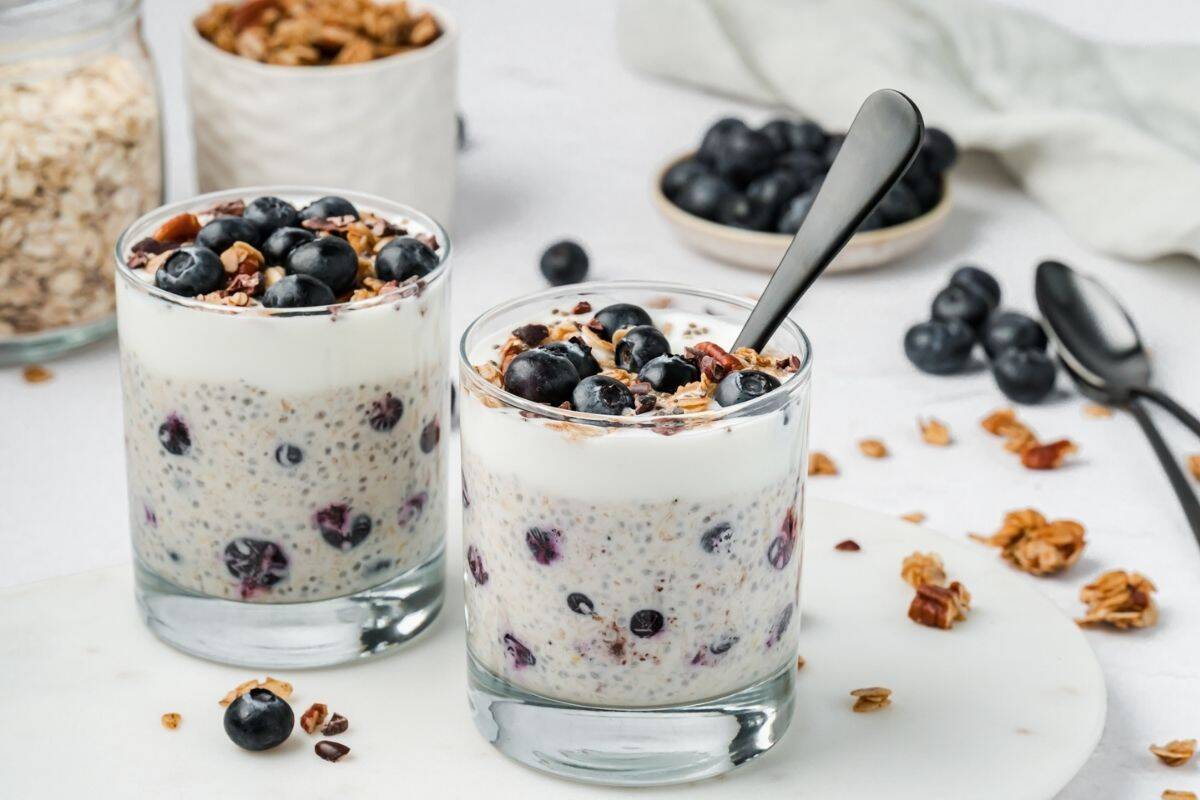 Two cups of blueberry overnight oats on a plate topped with granola, yogurt and fresh blueberries with a spoon ready for serving.