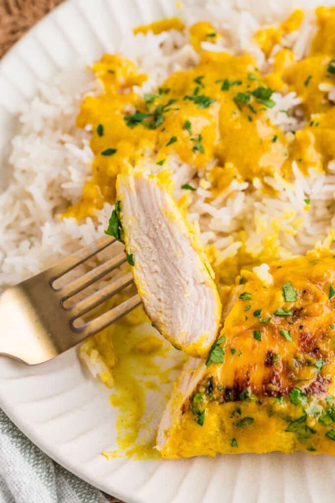 A slice of braised coconut chicken breast on a fork while plated with steamed rice.