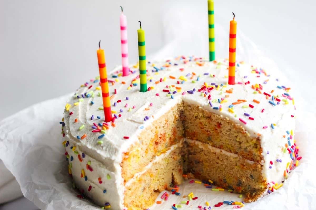 A freshly frosted and decorated gluten and dairy free birthday cake with candles that have just been blown out and a slice of cake removed for serving.