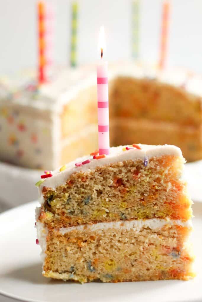 A slice of gluten and dairy free birthday cake being served on a plate with a lit candle on top.